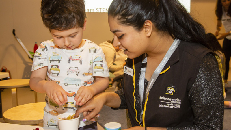 A woman with black hair in a ponytail sitting at a work table with a young boy with brown hair looking into a white cup full of things to create sound