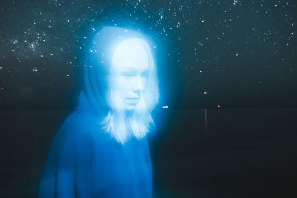 A woman looking pensive wearing a blue hoodie. There is a glow around her with stars in the background