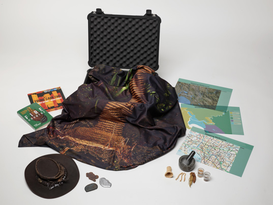 Museums Victoria Outreach Program Securing Food Futures learning kit containing the book Fire Country, the book Young Dark Emu, three samples of melted metal, a model eel trap with three model eels, a murrnong root, a mortar and pestle, transparent maps of Melbourne which can be overlaid with each other, a leather hat, a large banner with an image of the eel trap from Bunjilaka on it, and two seed samples (Kangaroo Grass and wheat)