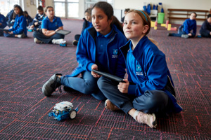 Two children sit behind a small robotic vehicle. Other children are seated in the background.