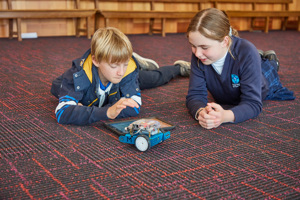 Two children stretched out on the floor behind a small robotic vehicle. One has a hand hovering above an iPad.