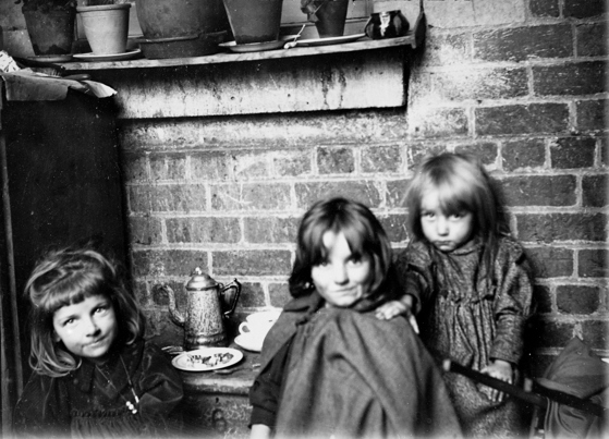 Three small girls having a tea party. There is the wall of a house behind them, and there are pot plants on the window ledge.