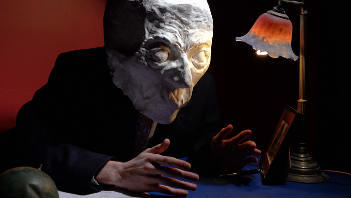 A person sat down at a desk wearing a suit and an oversized paper mache head.