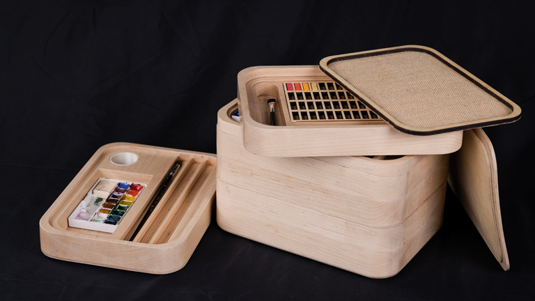 A rectangular wooden box with stackable layers, which contain paints and paintbrushes.