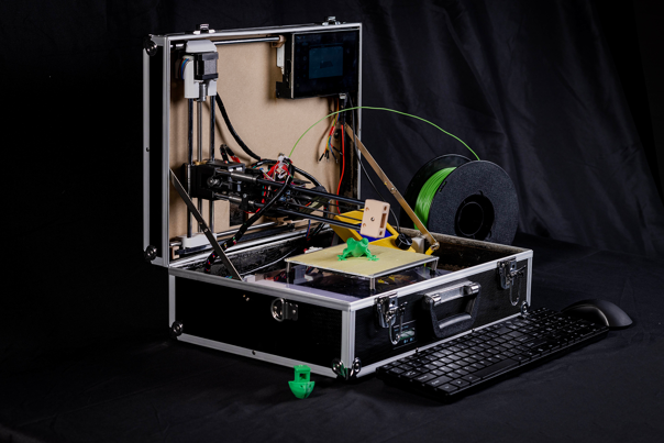 A photo of an open black suitcase containing 3D printer equipment and wiring, and two small green 3D printed models. A black keyboard and a black mouse sit in front of the suitcase.