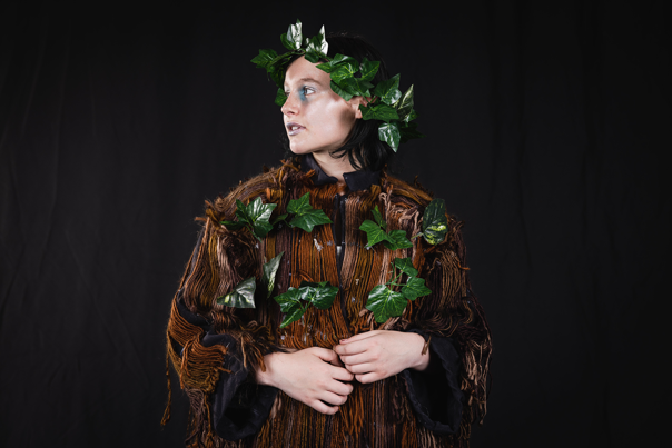 A person wearing a garment covered in brown thread and leaves, with a halo of leaves on their head. They are looking to the left of the frame.