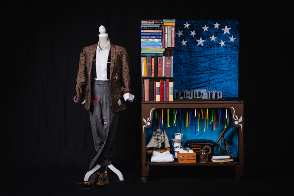 A mannequin dressed in a paisley patterned suit jacket, white shirt, braces and grey trousers. Next to the mannequin is a desk with numerous books and kick knacks placed on top of it.
