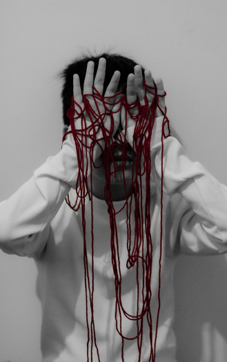 A black and white photograph of a person covering their face with their hands, with entangled red strings over both their palms.