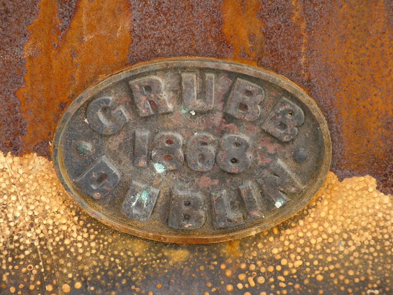 An oval metal plate, against a rusty and distressed metal background, bearing the words "Grubb 1868 Dublin"