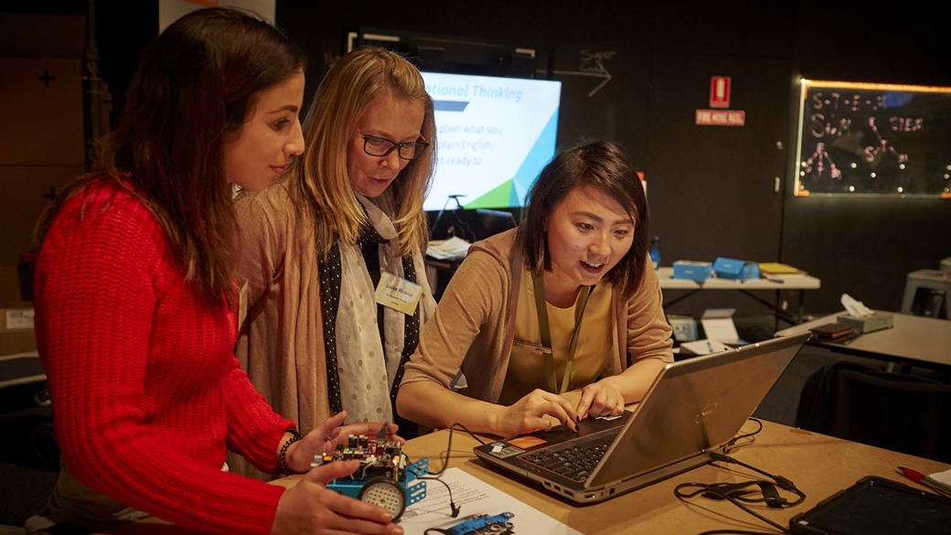 Two female teachers look on as a female museum staff member uses the mouse pad on a laptop. One teacher looks down at a blue car shaped robot with electronics on top.