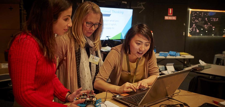 Two female teachers look on as a female museum staff member uses the mouse pad on a laptop. One teacher looks down at a blue car shaped robot with electronics on top.