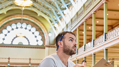 A man with a pen behind his ear sits on a chair sketching the ornate features of the Royal Exhibition Building interior.
