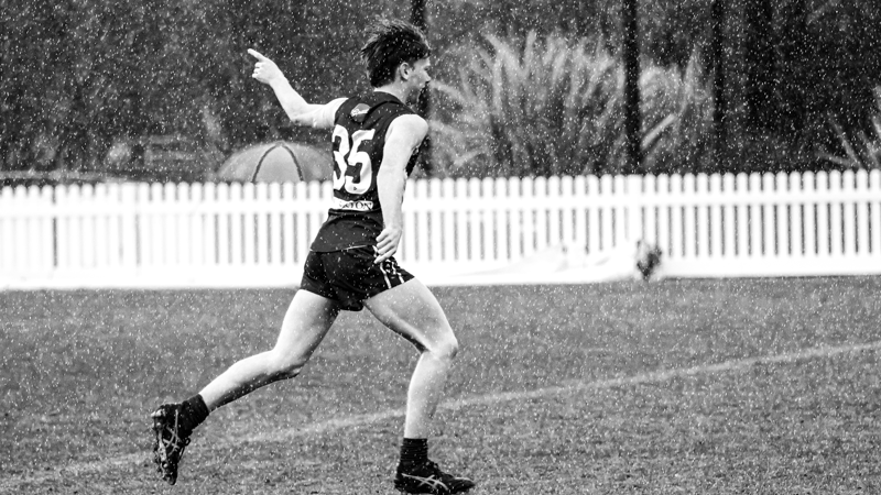 Black and white photo of young man in AFL uniform, running across a football field in the rain as he raises an index finger to the sky.
