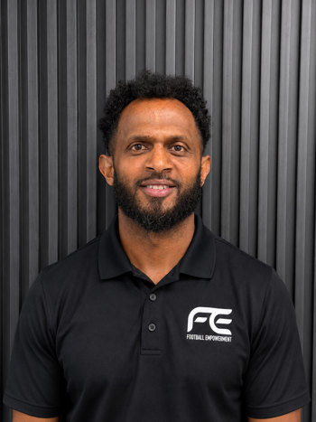 On a grey background, Tom Yabio smiles at the camera. Tom wears a black polo shirt with the logo which says Football Empowerment. 