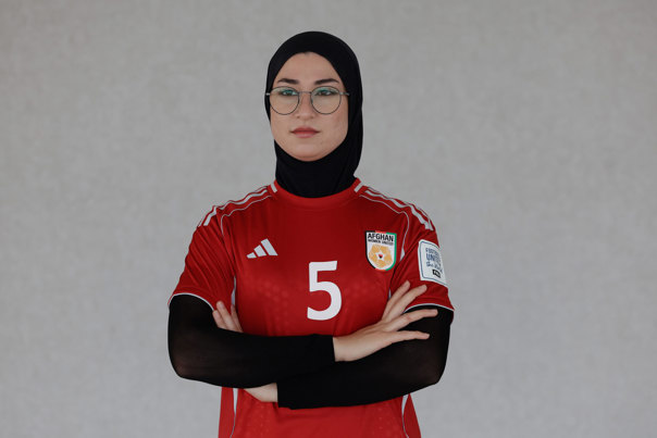 On a grey background, Mursal stands with her hands crossed. Mursal wears an Afghanistan Women’s National Team uniform with the number five and a logo on the T-shirt, black hijab and black long sleeves. The logo on the T-shirt says Afghan Women United. Mursal wears black round glasses.