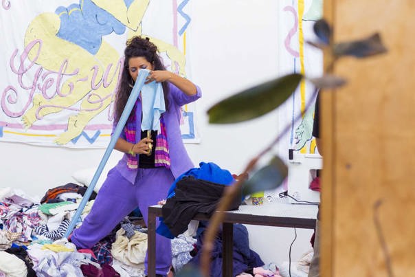 An individual in bright purple clothing snips a piece of blue fabric, in front of a pile of clothing waste. 