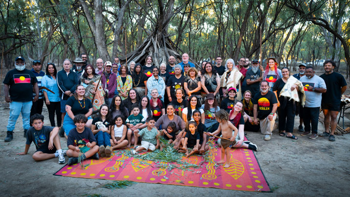 A large group of smiling people pose for a photo in front of a forested area. 