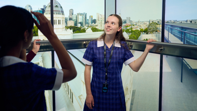 A student in the Road to Zero Experience Space standing in front of screens that simulate a glass elevator with the Royal Exhibition Building and Melbourne Museum visible on the screens 