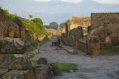 Looking down at a cobblestoned Pompeii street, which is framed by dwellings on the right hand side and a wall with greenery on the left