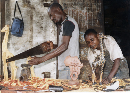 Two sculptors in a woodwork workshop. One man is standing using a saw to the left of the workbench, and the other man is working on fine detail of a carved giraffe sculpture, to the right. The workbench has an array of tools and sculptures in progress surrounded by wood shavings.