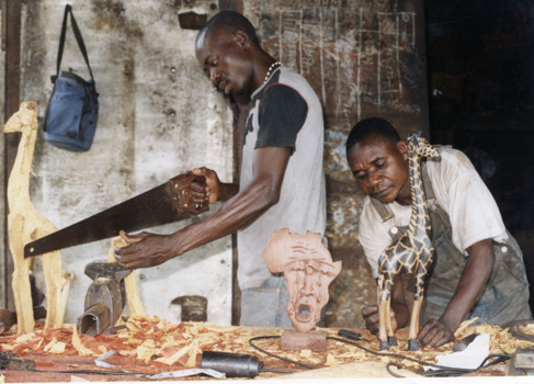 Two sculptors in a woodwork workshop. One man is standing using a saw to the left of the workbench, and the other man is working on fine detail of a carved giraffe sculpture, to the right. The workbench has an array of tools and sculptures in progress surrounded by wood shavings.