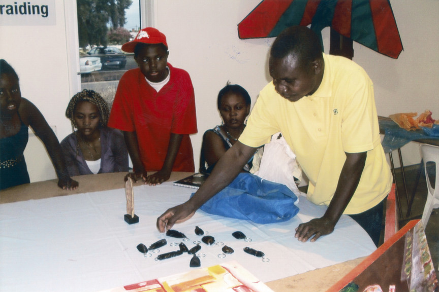 Nickel Mundabi  at the Shepparton Cultural Festival, arranging twelve carved keyrings on a table, as four people look on. Paintings and other artworks in the background.