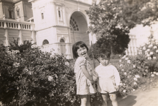 Two children near rose bushes outside partially visible building.