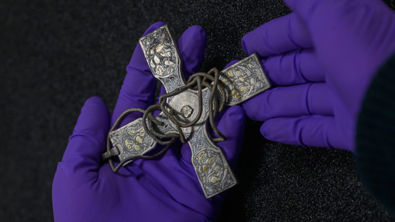 A close-up photograph of a person’s hands, wearing purple gloves, holding an artefact from the Galloway Hoard. The artefact is known as a pectoral cross. It is silver cross on a fine silver wire chain that would have been worn around the neck. Each of the four arms of the cross is decorated with symbols from the Christian faith.