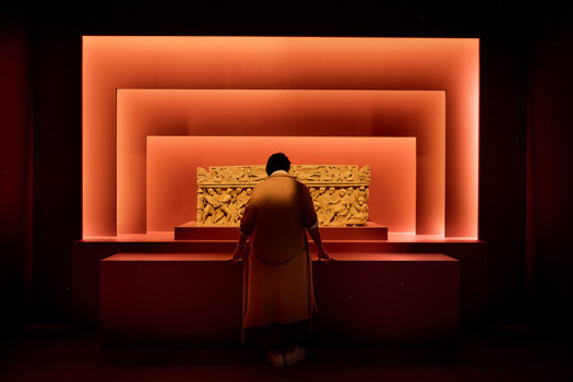 Person viewing an illuminated Roman sarcophagus framed by layered red exhibition walls. 