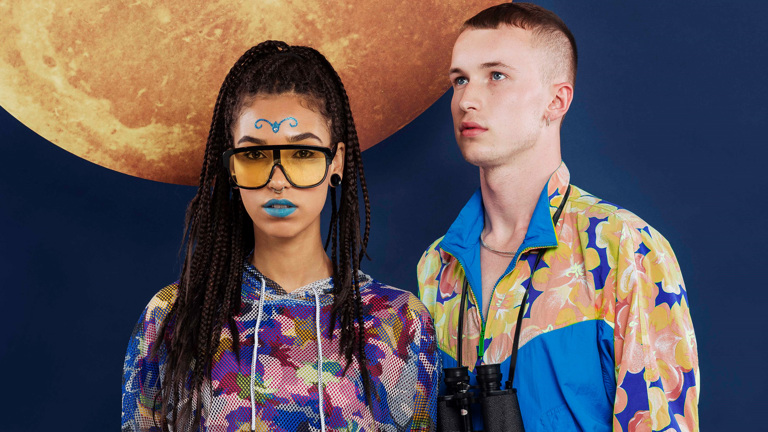 A young woman and man, each dressed in vivid colours, standing in front of a picture of a celestial body. She holds a thermos; he, binoculars.