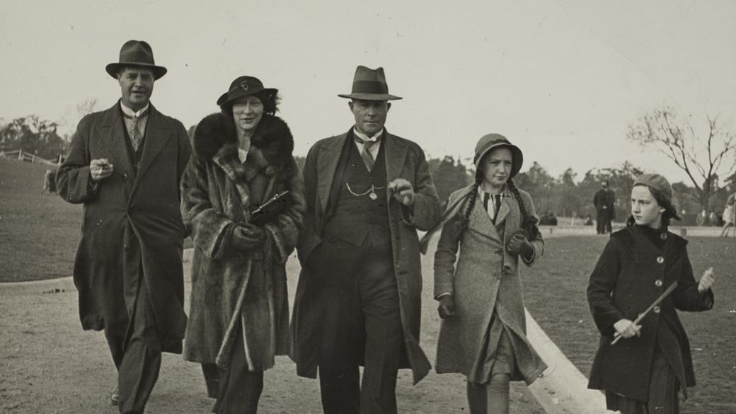 Two men, one woman and two girls in winter coats walking along a gravel path in a park. All are wearing hats.
