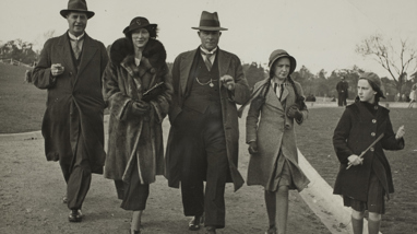 Two men, one woman and two girls in winter coats walking along a gravel path in a park. All are wearing hats.