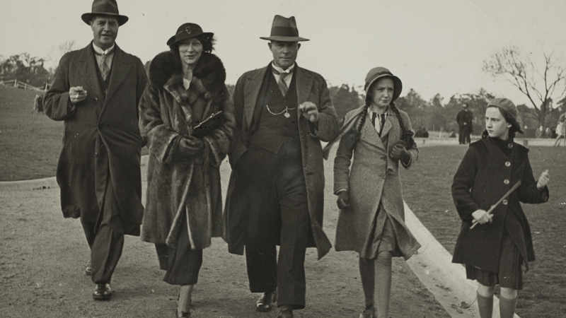 Two men, one woman and two girls in winter coats walking along a gravel path in a park. All are wearing hats.
