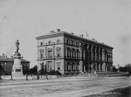A view of the Treasury Building, Spring Street, Melbourne, showing the front entrance and surrounding gardens. A sculpture of a man is located to the left of the building, and a British flag is flying from the roof. 
