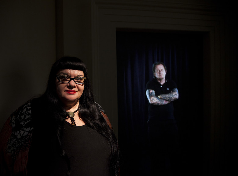 Woman who is in shadow standing in front of a screen displaying a man with tattoos on his arms