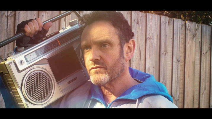 Film still featuring a man in a blue hoodie, holding a vintage boombox on his shoulder, against the backdrop of a wood fence.