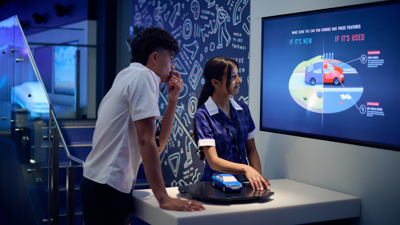 Two students in the Road to Zero Experience Space standing in front of a screen with a blue model car in front of them 