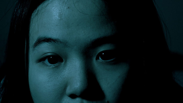Film still showing a close-up of a young woman's face lit in teal-blue tones against a dark background, with dramatic shadows emphasising her features.