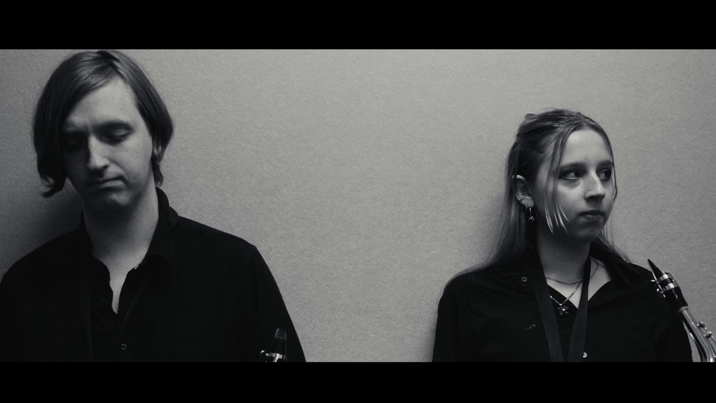 Black-and-white image of two musicians in dark clothing, each holding wind instruments, standing against a plain wall with solemn expressions.