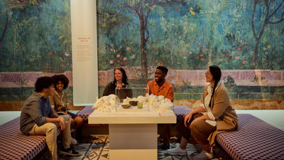Group of visitors seated around a table exhibit, with a painted Roman garden mural filling the background in ROME: Empire, Power, People exhibition in Melbourne Museum.. 