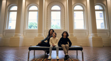 A woman, a girl and a boy sitting on a bench in the Long Room at the Immigration Museum
