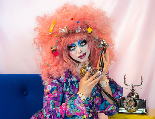 Drag performer Belial B'Zarr with curly pastel pink hair and a brightly patterned purple suit. They are sitting on a blue chair and holding a vintage telephone receiver.