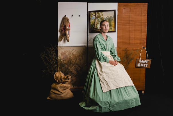 Woman in long green dress with a white apron, sitting in front of a wooden backdrop with a framed painting, hanging animal carcass, dried plants, and a "Staff Only" sign.