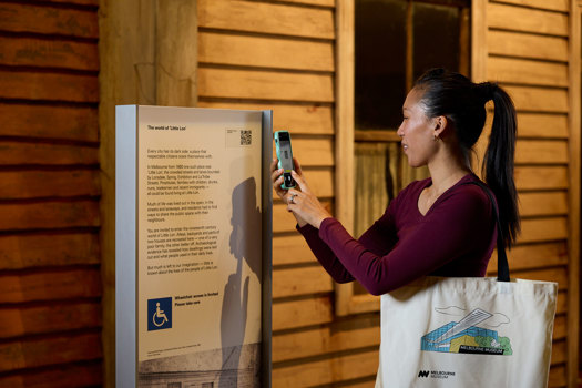A woman scans a QR code next to the Little Lon exhibition showcasing an early home from the area.