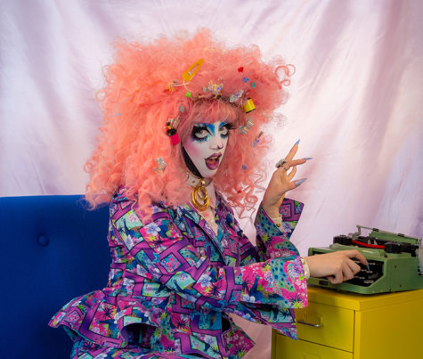 Drag performer Belial B'Zarr with curly pastel pink hair and a brightly patterned purple suit. They are sitting on a blue chair and posing with a vintage green typewriter.