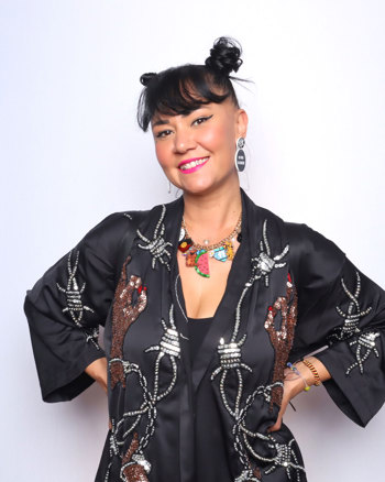 Woman looking directly at the camara. She has dark hair, dark eyeliner and pink lipstick. She is wearing a chunky necklace and a black jacket with beaded with barbwire and arms motifs..