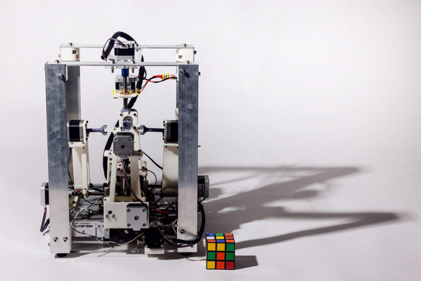 Aluminium frame device with exposed motors, articulated arms, and visible wiring, with a Rubik's Cube positioned at its base.