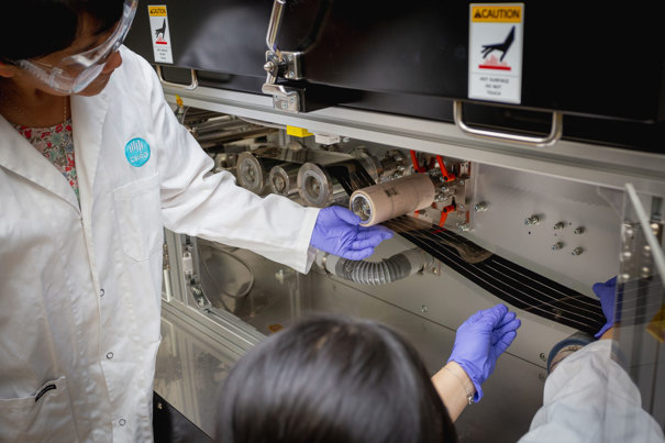 Two people in lab coats, gloves and safety glasses guiding a film through a machine