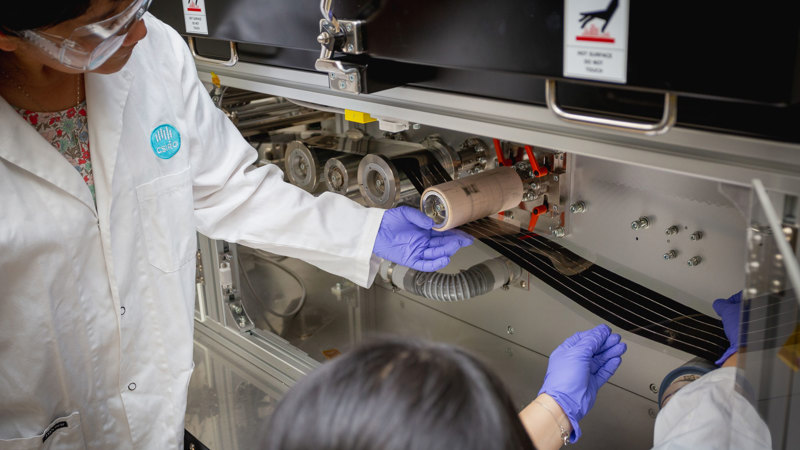 Two people in lab coats, gloves and safety glasses guiding a film through a machine