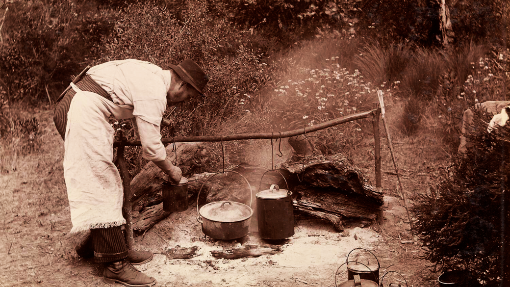 A member of the Field Naturalists Club of Victoria cooking at a camp fire, preparing meals for the men during the Club's scientific expedition to the Furneaux Group of islands in November 1893. There are two billy cans and a cooking pot over the fire. Three other billy cans and a pail are on the ground to the right.